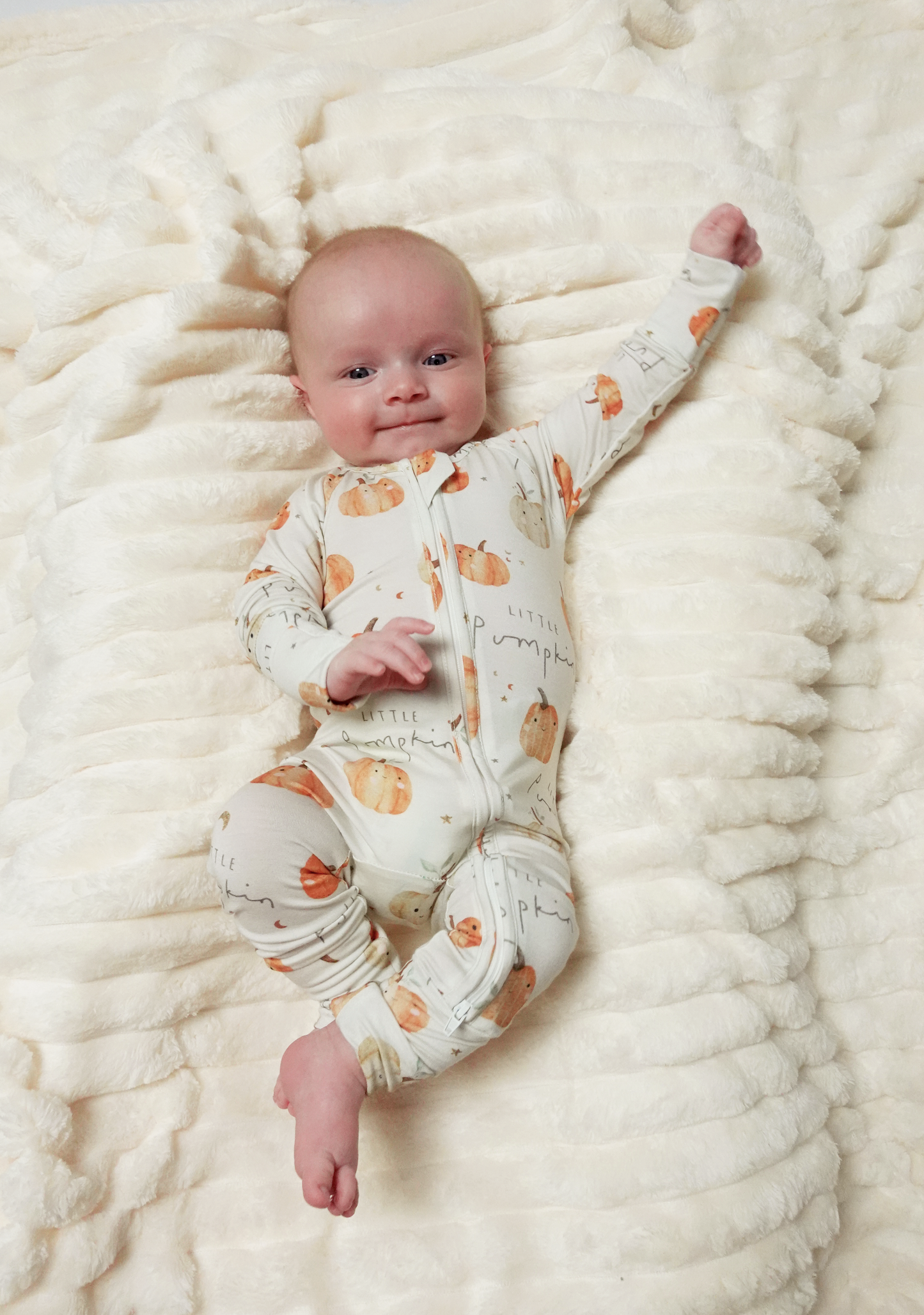 A baby lying on a textured cream blanket, wearing a cream-colored footed sleeper with an orange pumpkin and leaf pattern.