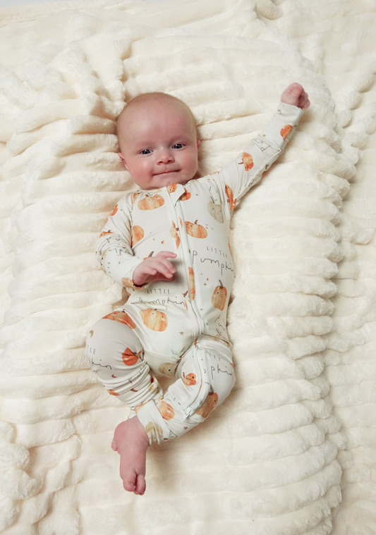 A baby lying on a textured cream blanket, wearing a cream-colored footed sleeper with an orange pumpkin and leaf pattern.