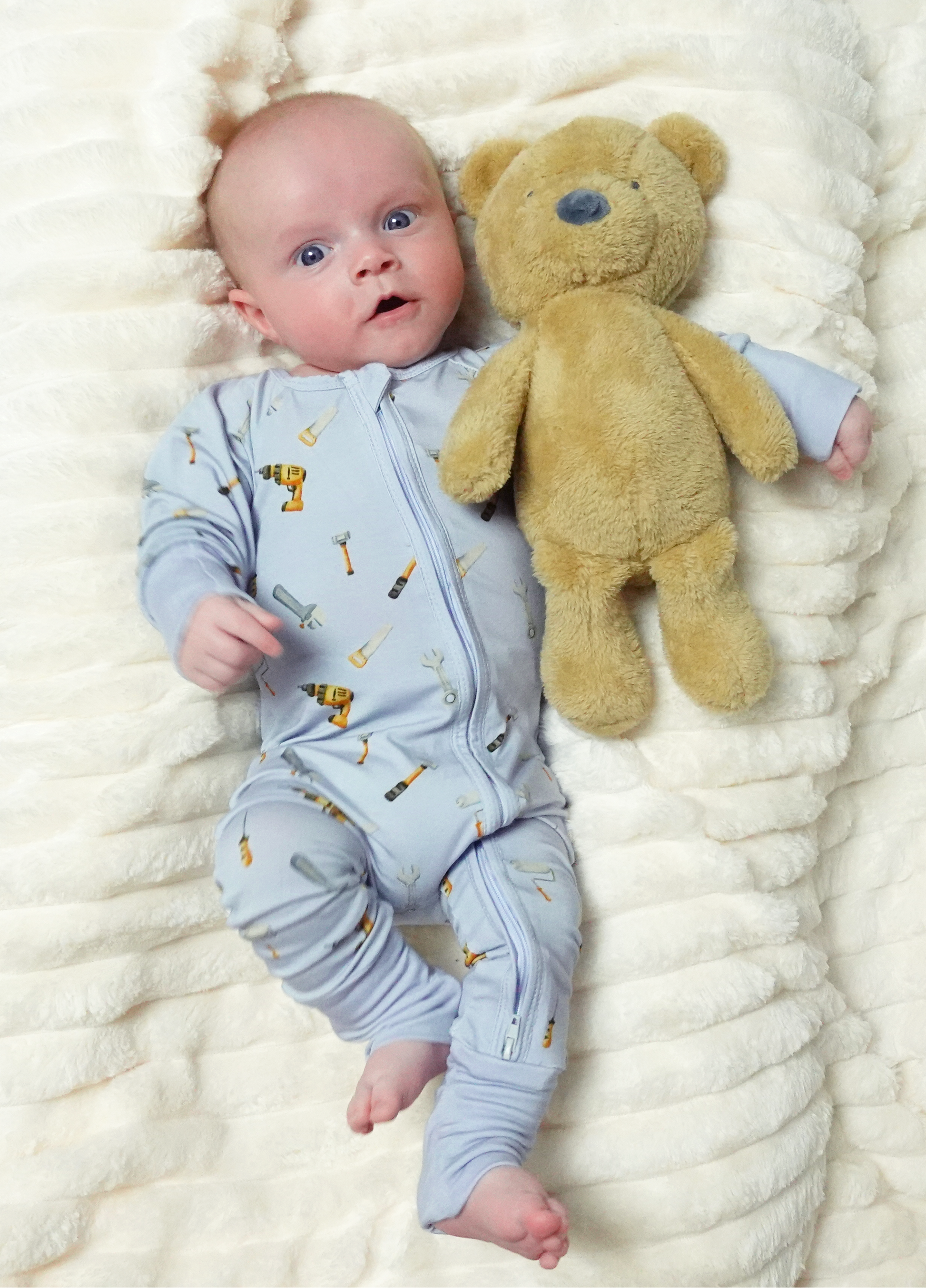 A baby lying on a textured cream blanket next to a plush teddy bear, wearing a light blue one-piece jumpsuit with a tool print.
